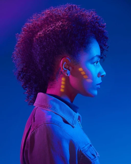 Side profile portrait of a woman with afro hairstyle, geometric makeup, wearing a jacket, illuminated by pink and blue lighting.